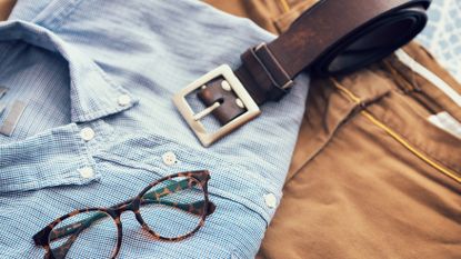One of the best belts for men laying on top of an outfit consisting of a blue shirt, brown chinos, and glasses