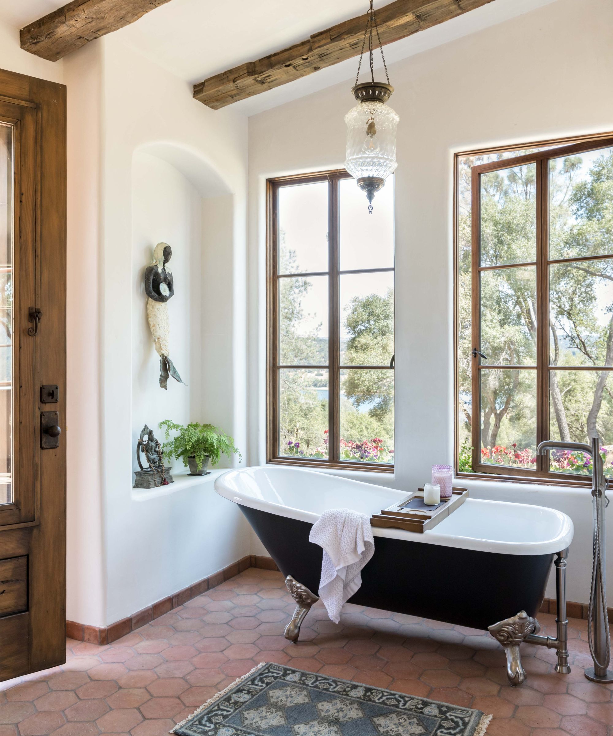 A Mediterranean bathroom with terracotta floor tiles, plaster walls, exposed wooden ceiling beams, and a black freestanding bath