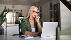 An older woman looks serious as she works on her laptop at a table at home.