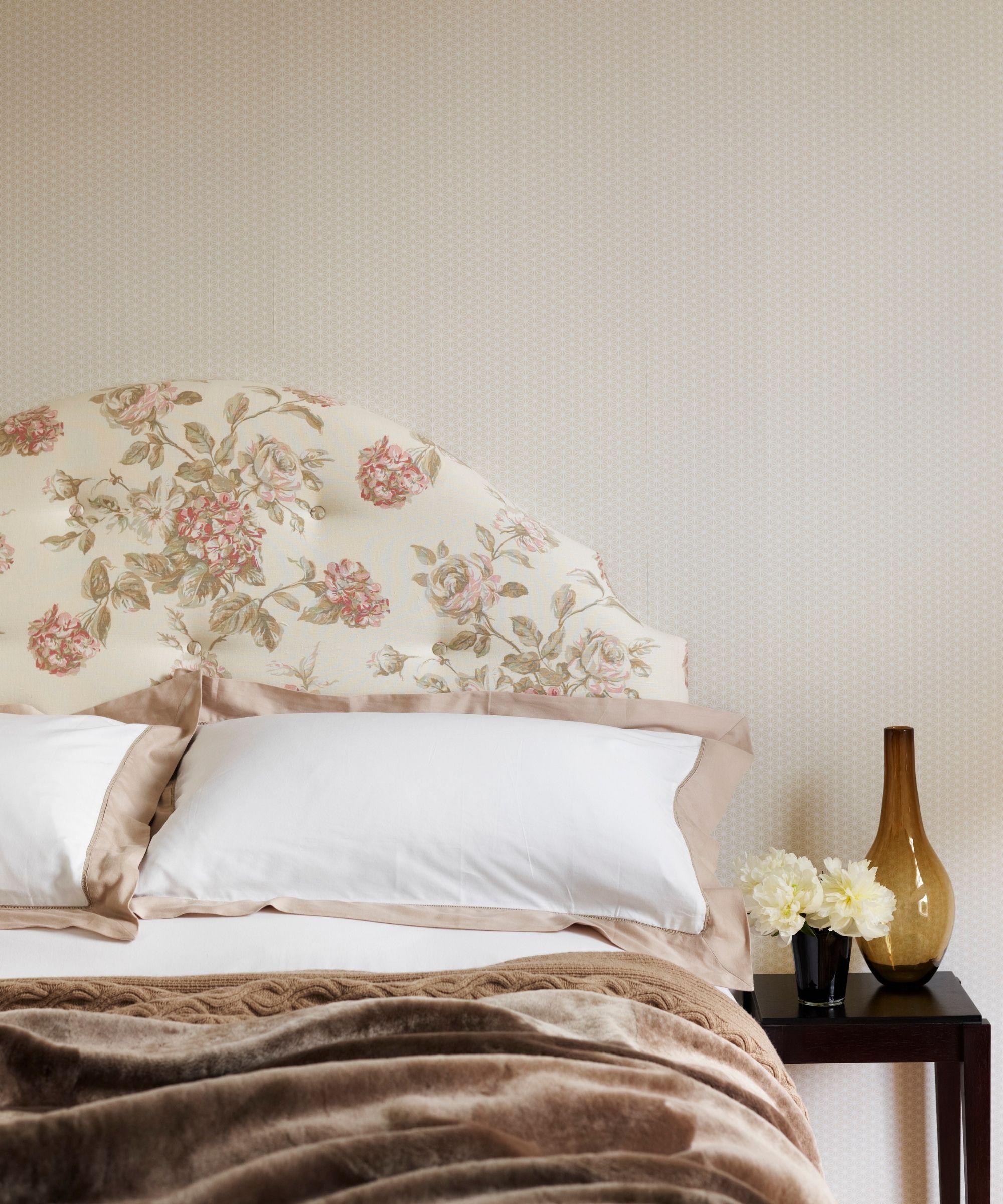 A soft pink bedroom with pale wallpaper, a white floral headboard, white and peach bedding, and a brown blanket. Beside the bed is a black bedside table with white flowers and a brown glass vase.