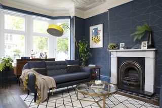 Blue living room with blue and gold geometric wallpaper, white fireplace, blue sofa, yellow floor lamp, black and white rug and brass and glass coffee table