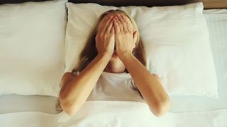 Blonde woman lies in bed with hands covering her face after experiencing a nightmare