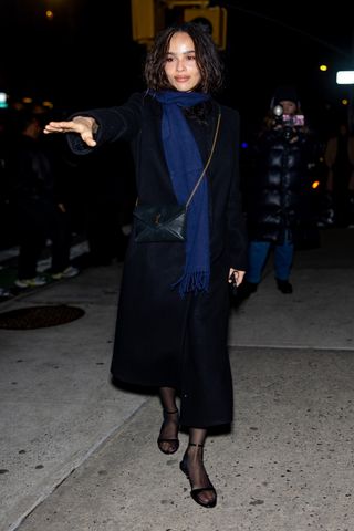 Zoe Kravitz wears a blue scarf, long black coat, ysl bag, black sheer tights, and black open-toe sandals.