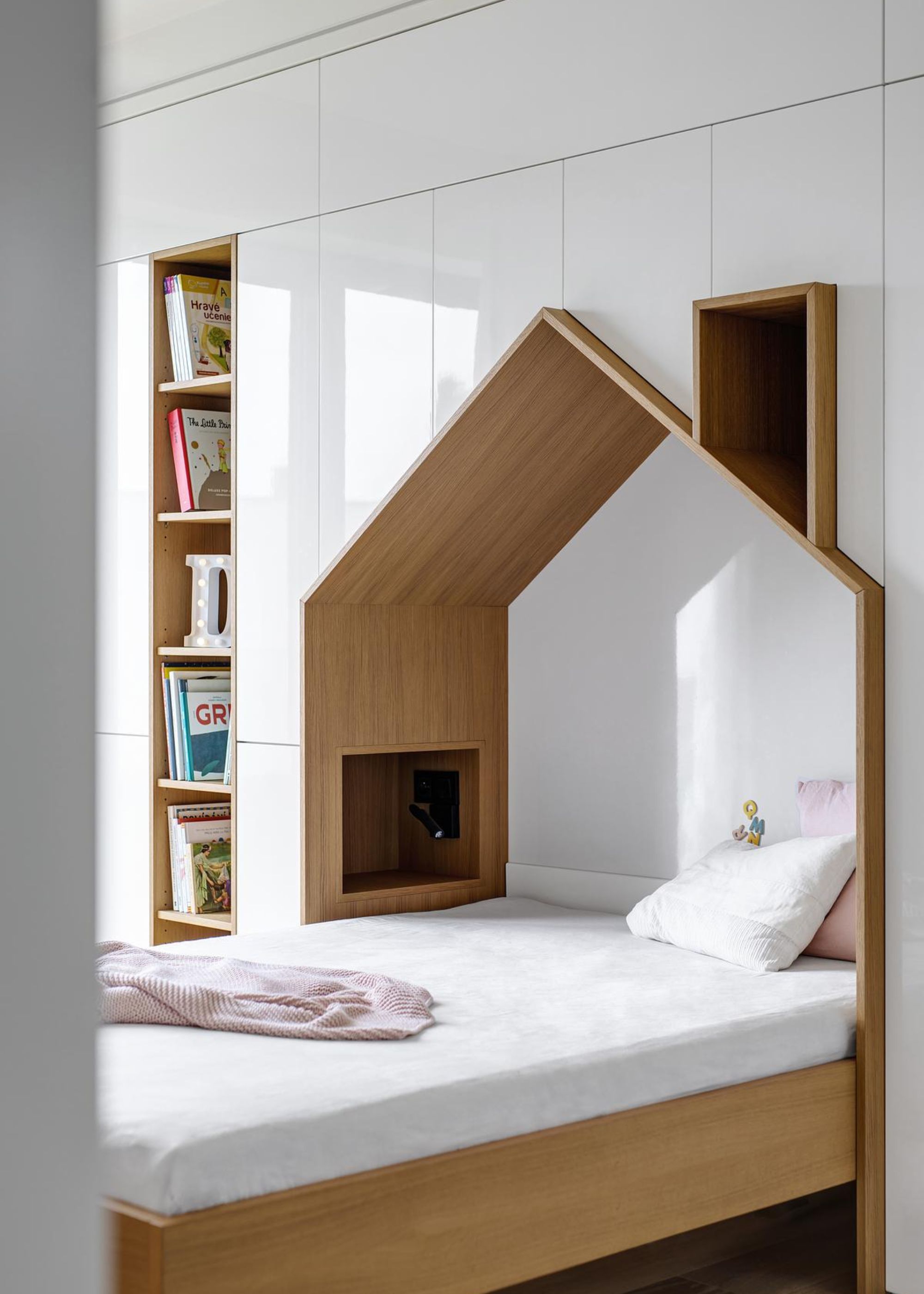 A bedroom with a bed that has one pillow on top of it and a triangular curves headboard as well as a view of the bookshelves next to the bed