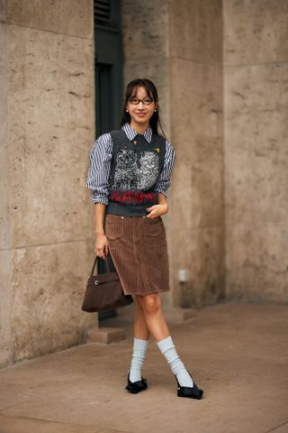 Woman at fashion week wearing preppy outfit featuring a blue striped button-down, cat print Ganni sweater vest, brown skirt, socks, and black shoes.