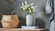 IKEA FL&Aring;DIS Basket against a dark grey wall beside a stack of magazines with a white fluted vase filled with white flowers.