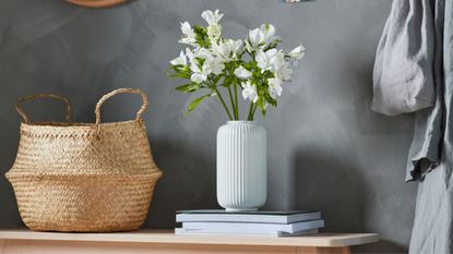 IKEA FL&Aring;DIS Basket against a dark grey wall beside a stack of magazines with a white fluted vase filled with white flowers.