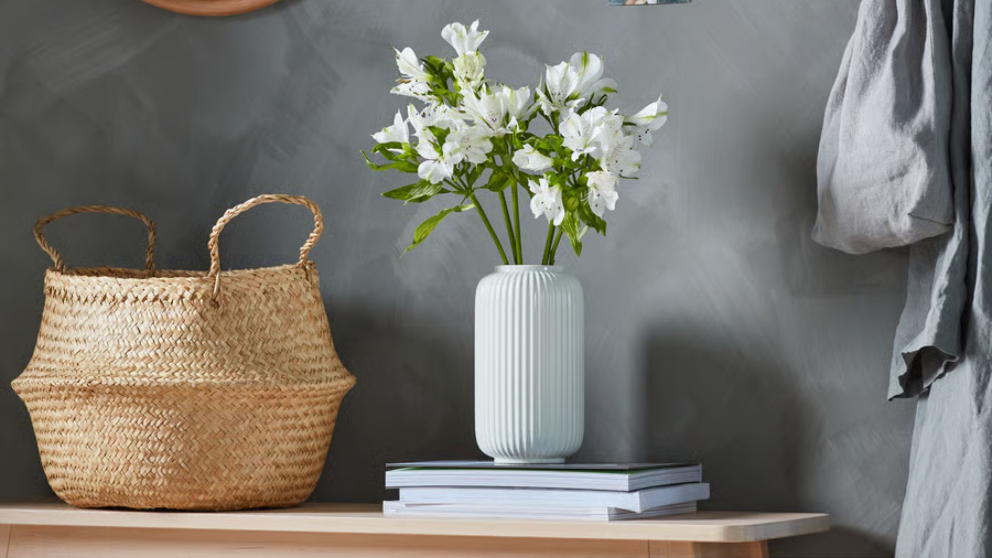 IKEA FL&Aring;DIS Basket against a dark grey wall beside a stack of magazines with a white fluted vase filled with white flowers.