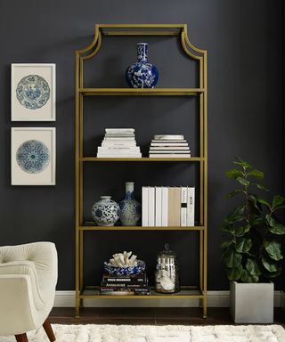 Moody dark wall, with shapely brass open bookshelf, styled with white and deep blue books and chinoiserie style vases.