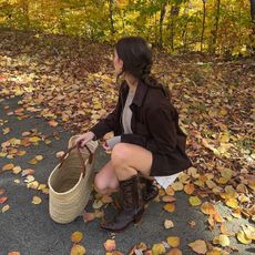 Woman wears brown cowboy boots with a brown jacket and basket bag on a leafy lane