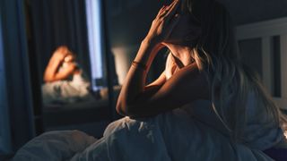 The image shows a blonde-haired woman sitting up in bed in the dark with her hands on her face
