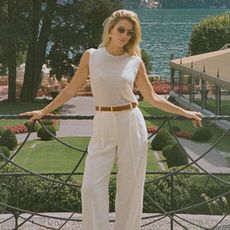 Woman standing in white muscle tee, white pants, brown belt, and sunglasses in Lake Como, Italy.