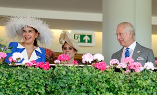 Sophie Winkleman in a white feathered hat and blue coat standing behind a floral railing next to Princess Beatrice and King Charles at Royal Ascot