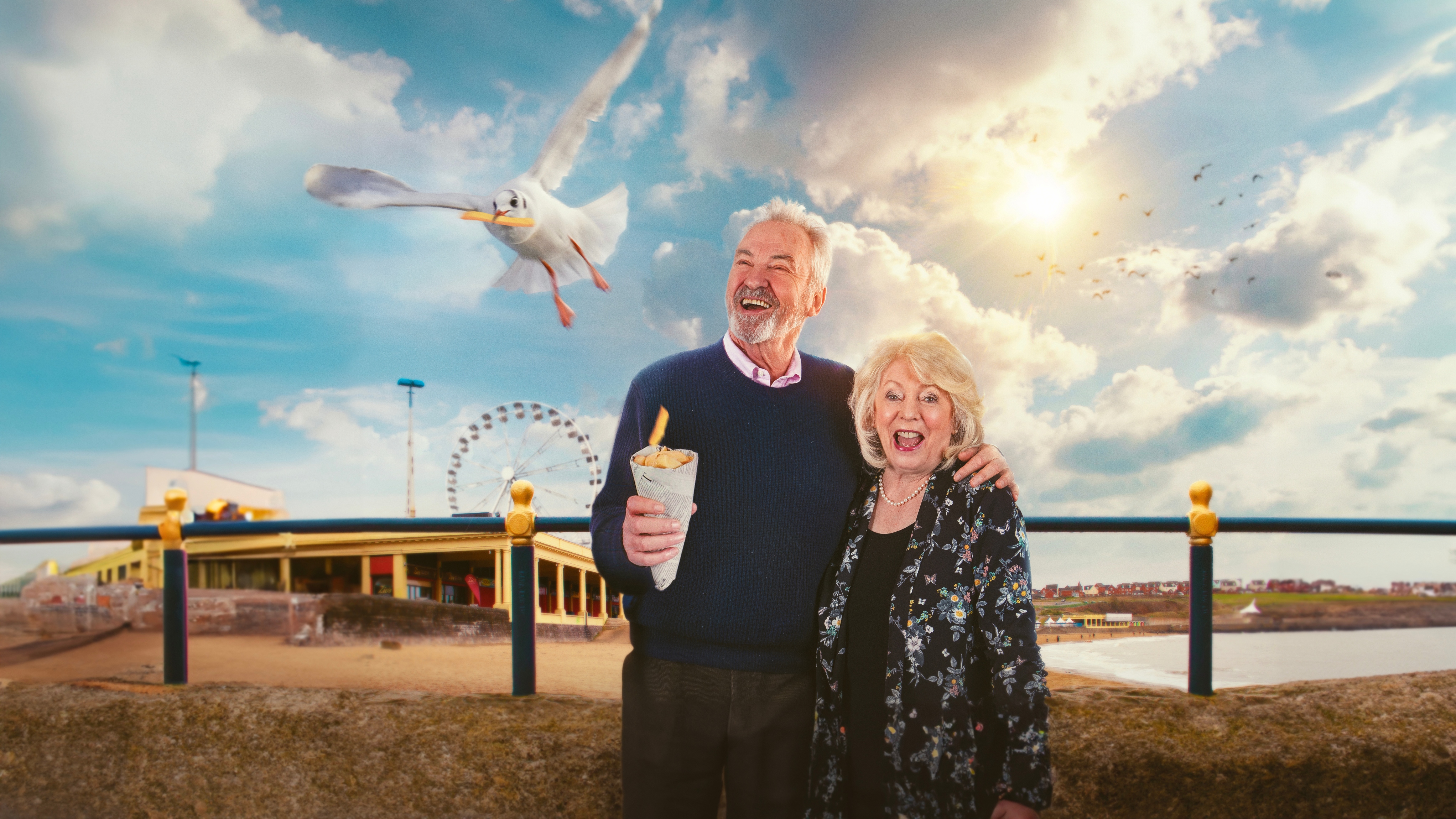 Alison and Larry holding chips with a seagull circling above them with the seaside in the background.