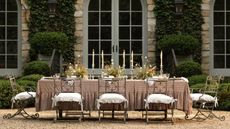 large long outdoor table styled on a shingle patio area with a pink plaid ruffle tablecloth, ornate chairs, laid with flowers and tall taper candles