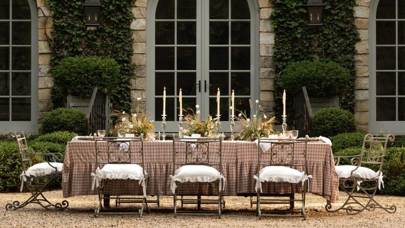 large long outdoor table styled on a shingle patio area with a pink plaid ruffle tablecloth, ornate chairs, laid with flowers and tall taper candles