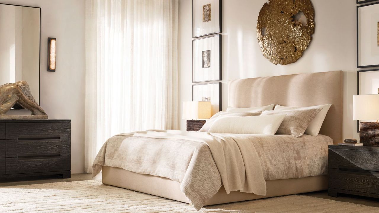 White and beige bedding on a beige bed frame against a white wall with gold wall art; to the left, a mirror hanging above a dark wood dresser; to the right, a lamp on a dark wood nightstand. 