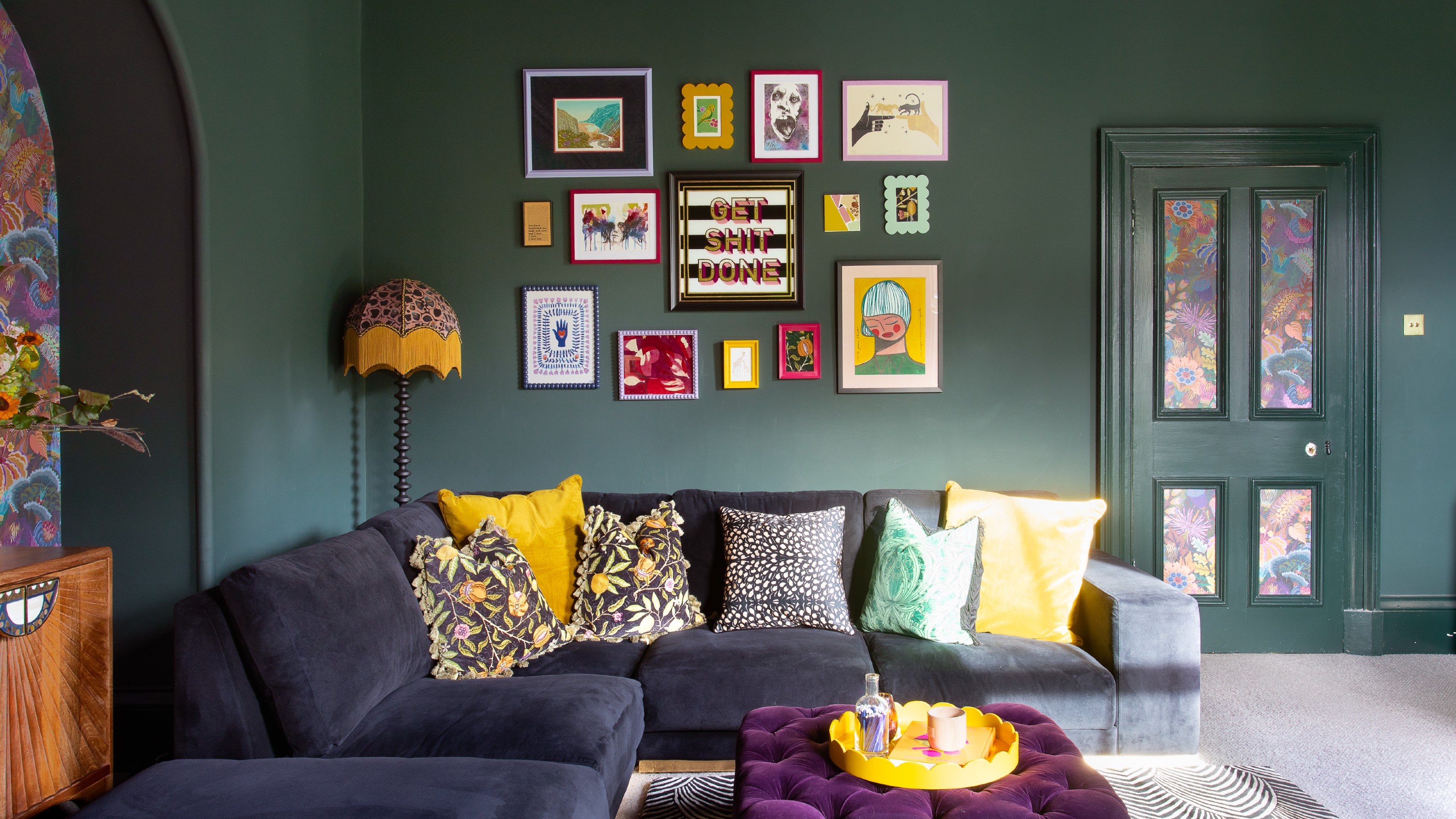 A living room painted in a dark green shade with a corner sofa in blue velvet and a colourful gallery wall hung above it 