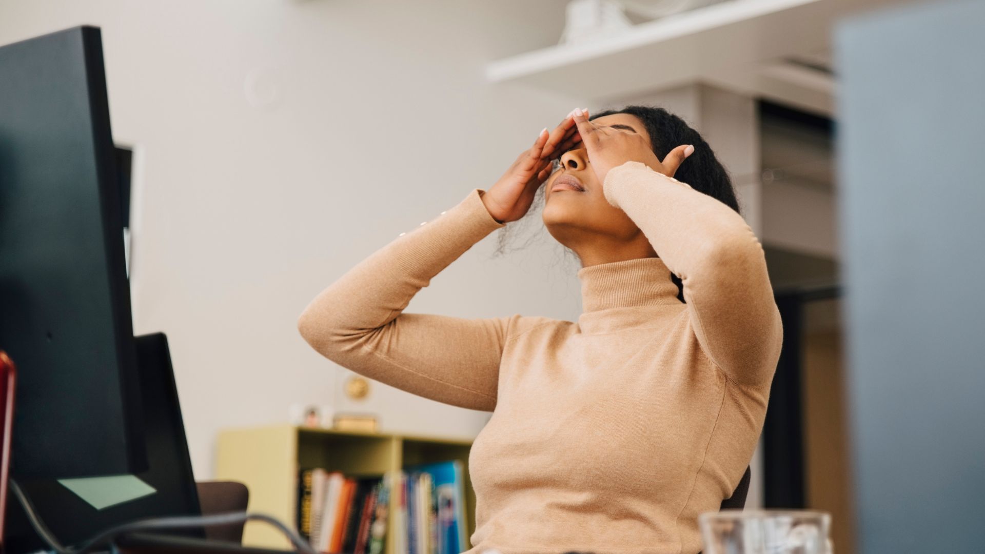 Woman holding hands over head, representing a headache at work desk