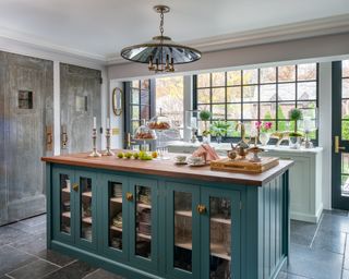 A kitchen with blue-green wooden island and brass and steel doors leading to a cupboard and powder room