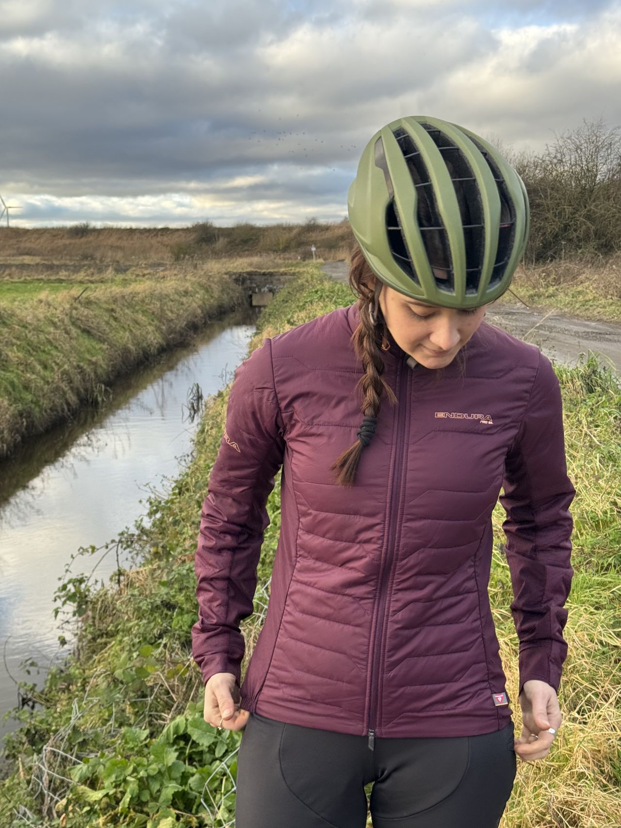 Woman in burgundy jacket, face looking down, with a green helmet and a scrubby gravel track behind.
