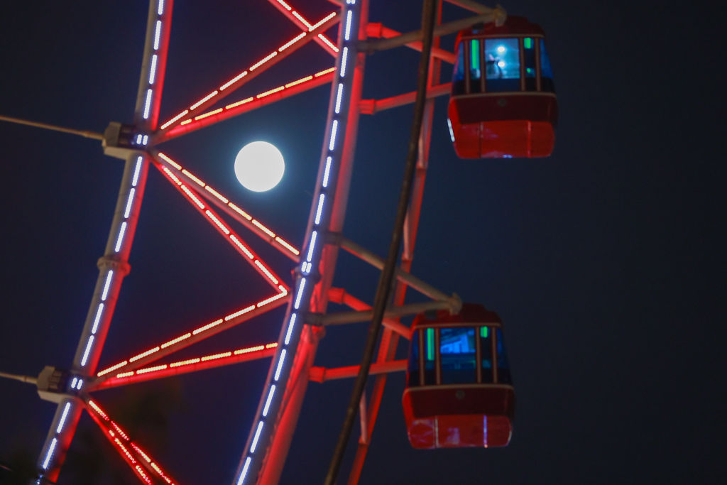 Close-up de uma roda gigante vermelha com luzes acesas e a lua brilhando atrás dela.
