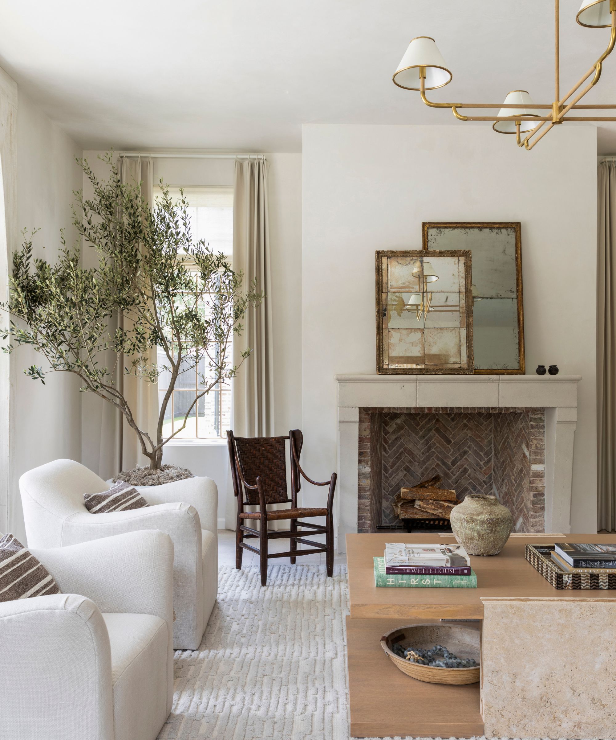 A light and airy transitional living room with soft white walls, a cream rug, two white armchairs, a vintage chair, a light wooden coffee table, and a fireplace with antique mirrors.