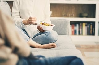 Person sat crossed legged on a grey sofa holding meal in fibre food packaging tray by Huhtamaki, blurred front and back surroundings