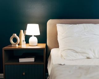 Bed with headboard and dark teal side table with white lamp glowing against against a teal wall