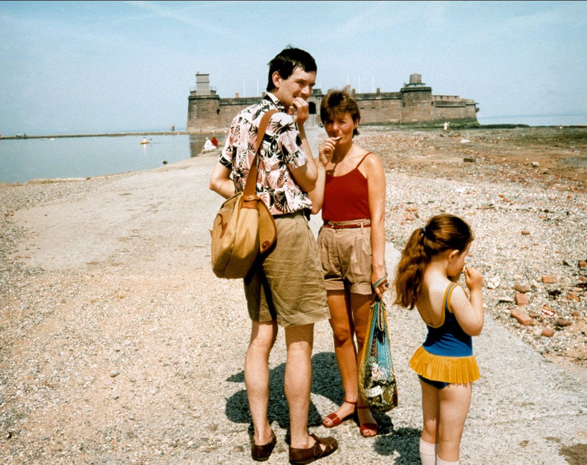 A young Martin Parr in a floral shirt and his wife Sue in a red top stand on a rocky beach near a young girl in a blue swimsuit, with a historic stone fort visible in the background.
