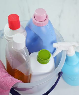 Bottle of bleach among other unmarked bottles of cleaning solutions in a clear plastic bucket on a neutral toned background