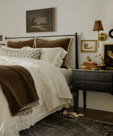 a rustic bedroom with a bed that has white and brown sheets on it, a small nightstand with decor on top including books, decorative objects, a vase, and a photo, and a wall sconce beside the bed