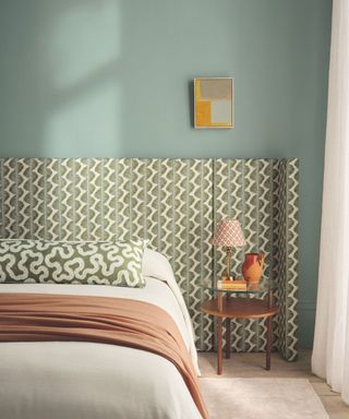 Printed headboard with chevron design, bed with pink and green throw and cushion and bedside table in wood with orange vase and printed lampshade