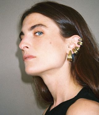 Maria Sole Ferragamo with her head tilted to the side whilst looking at the camera, wearing hair down, a black dress and modelling gold earrings, beige background.