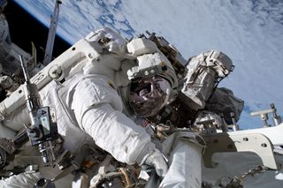 nicole mann in a spacesuit during a spacewalk, working on hardware on the international space station. behind is cloudy earth and black sky