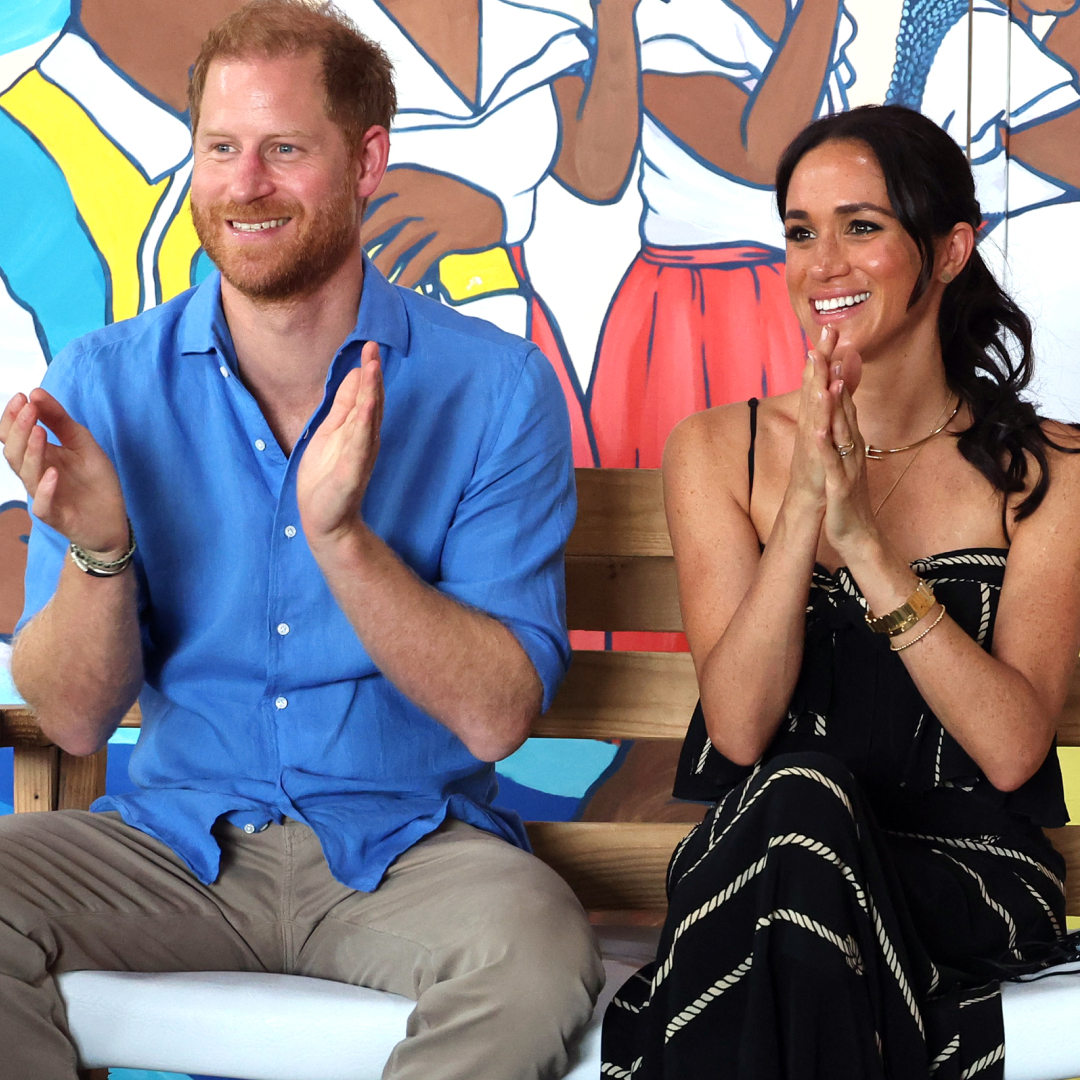 Prince Harry wears a blue shirt and brown pants and claps next to wife Meghan Markle who wears a black and white striped dress
