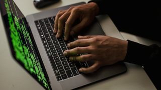 A hacker typing on a MacBook laptop with code on the screen.