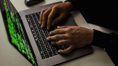 A hacker typing on a MacBook laptop with code on the screen.
