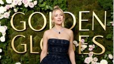 Kate Hudson in a black strapless dress standing in front of a green, moss and floral background that has the Golden Globes logo in large gold letters on it