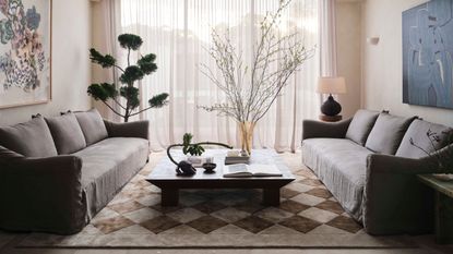 living room with linen sofas, check rug, square coffee table, bonsai tree and branches, and artwork on walls in front of sheer curtains