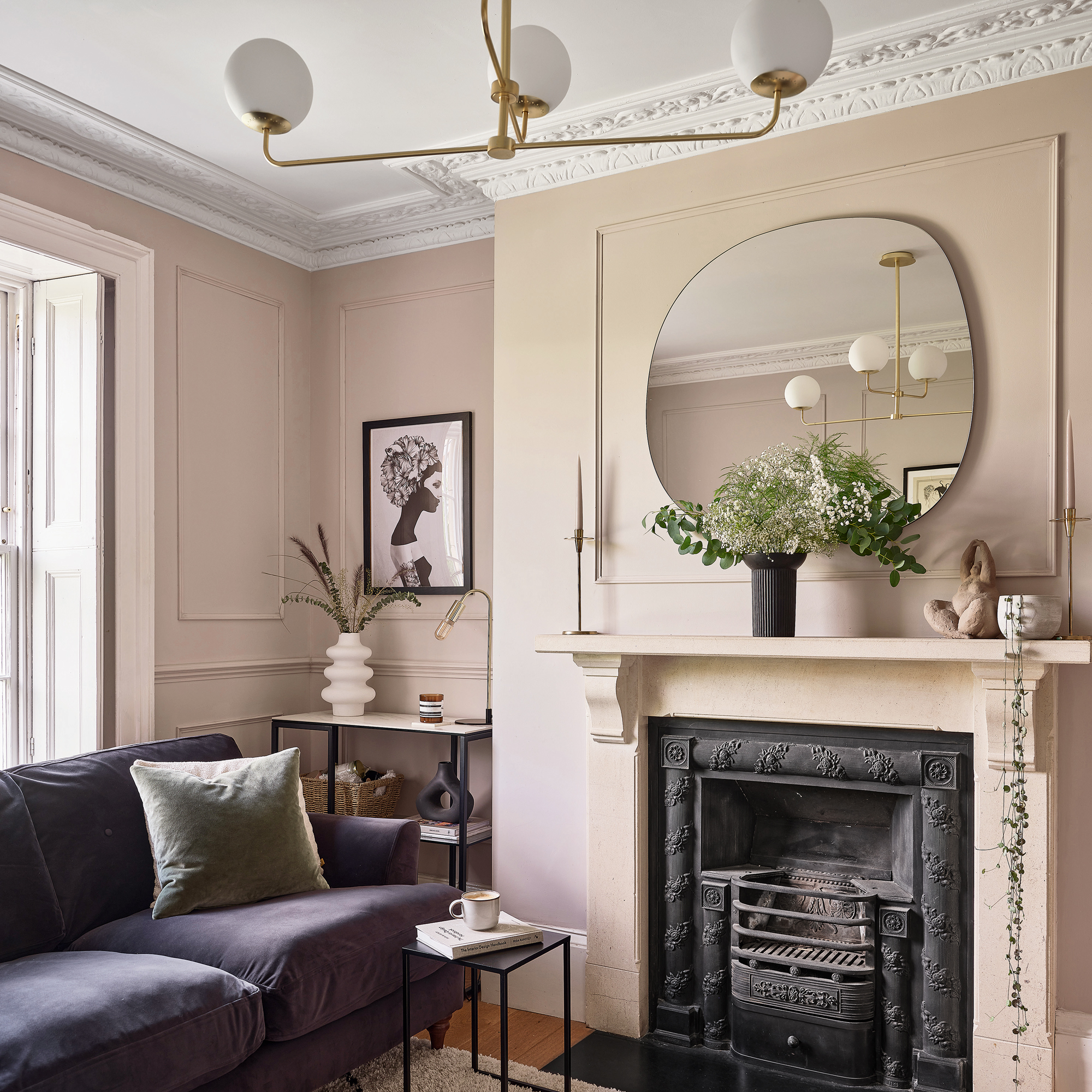 a pale pink living room with a dark purple velvet sofa fireplace, large circular overmantel mirror and classic cornicing