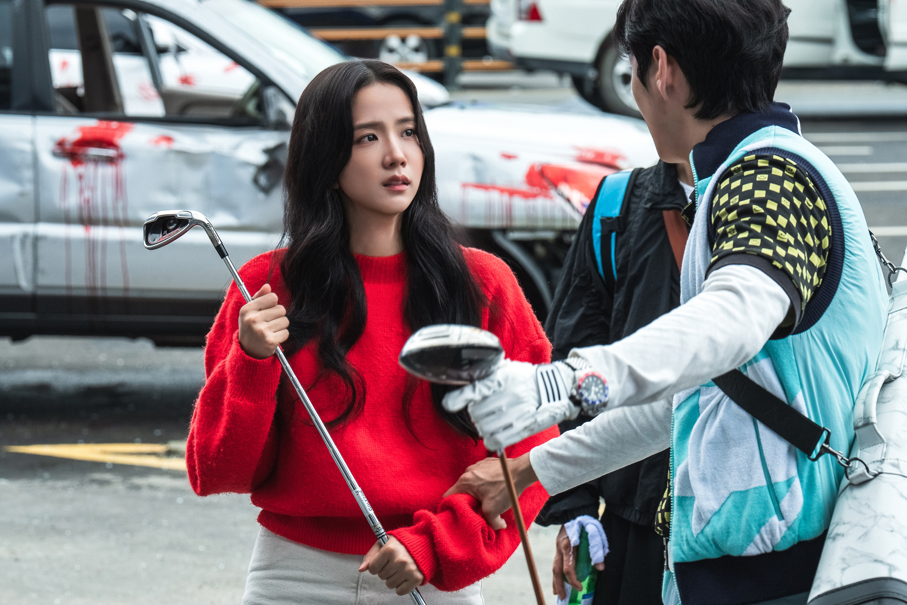 A woman (Jisoo of Blackpink) holds a golf club while standing with two unseen men also holding clubs, as the group stands outdoors in front of a white blood-splattered car. A still from the Prime Video K-drama 'Newtopia.'