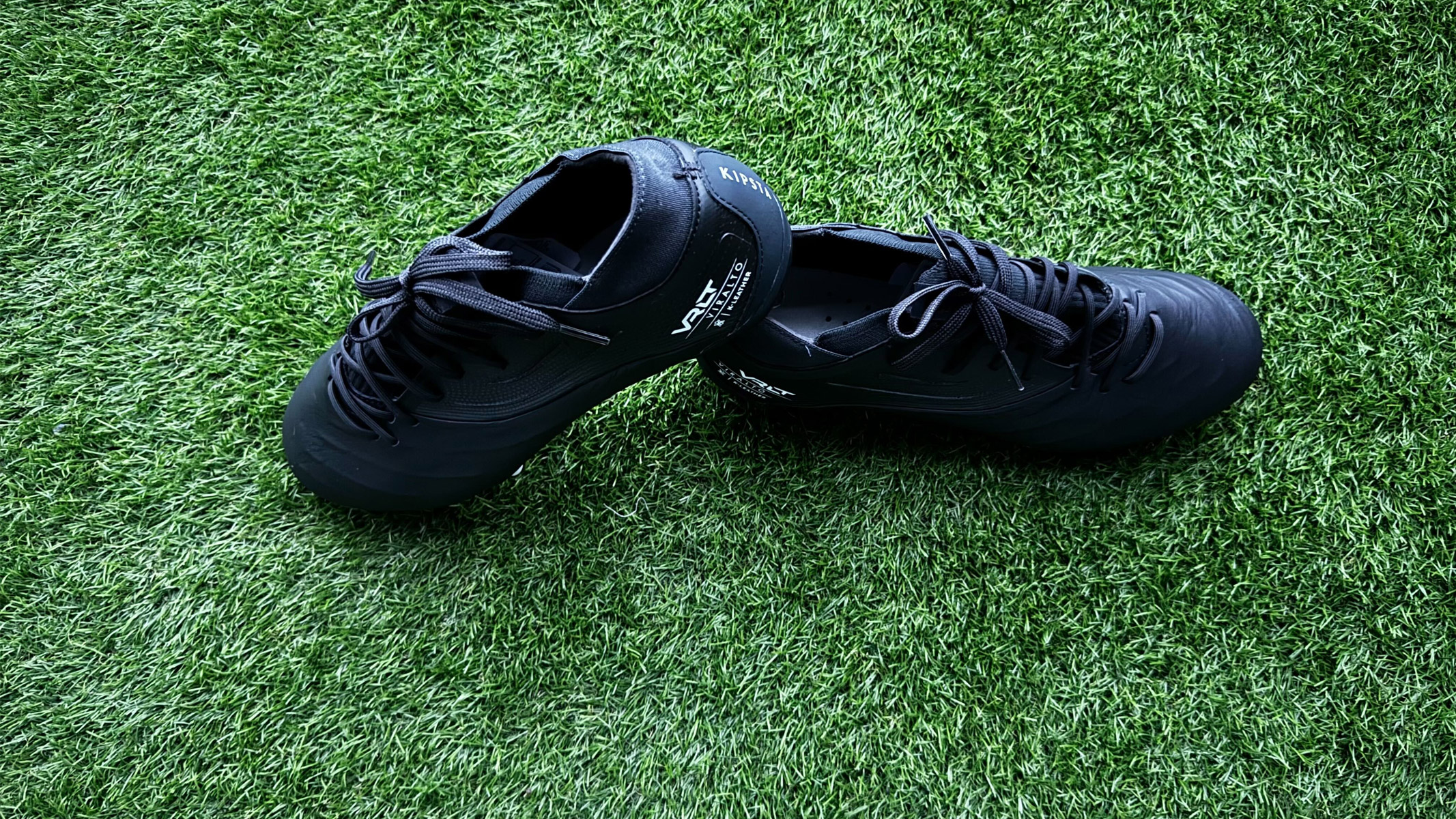 Two black football boots, one heel is resting on top of the other, are back to back on green artificial grass. Kipsta Viralto IV Pro