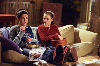 lauren graham and alexis bledel sit on the couch eating snacks in gilmore girls