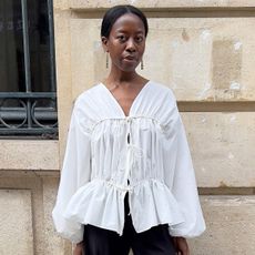 Influencer stands in front of Parisian brick wall wearing white frilly blouse and tailored black pants.
