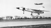 a jet plane landing on a runway accompanied by another plane behind it, in mid-air. the image is in black and white due to age. bare trees are visible in the background