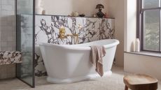 A white roll top bathtub against a large pattern marble half wall. A glass shower door separates a matching marble bench. Hold hard wear. A towel is draped over the edge of the bath. 