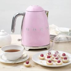 Pink Smeg kettle available at Boots, on a wooden table with a cup of tea and sweet treats. 