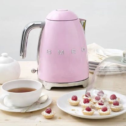 Pink Smeg kettle available at Boots, on a wooden table with a cup of tea and sweet treats. 