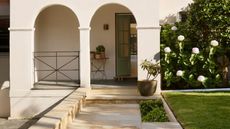 Image of the front of a white house that has two archways leading to a front stoop with a sage green door that is halfway open. There is a stone walkway and a grassy front yard